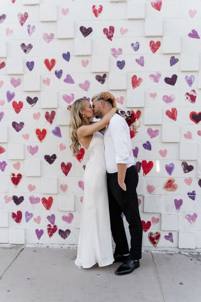 A bride and groom hold each other tightly and kiss as they pose for photos in front of a white brick wall painted with little colorful heart shapes.