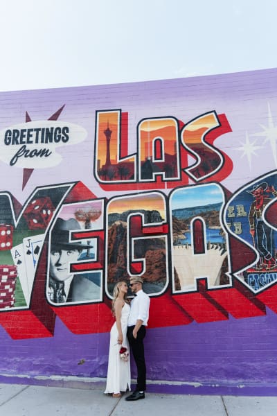 A bride and groom stand closely together and look at each other as they pose for wedding photos in front of a mural that looks like a postcard that says, "Greetings from Las Vegas".