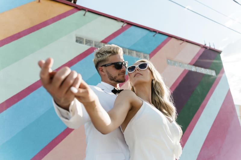 A low angle shot of a bride and groom posing in front of a large mural with colorful stripes. He stands behind her and they hold right hands straight out to the side. 