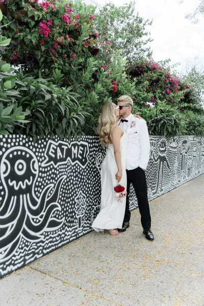 A bride and groom kiss as they stand in front of a raised flower bed. The wall of the flower bed is painted with a black and white mural.