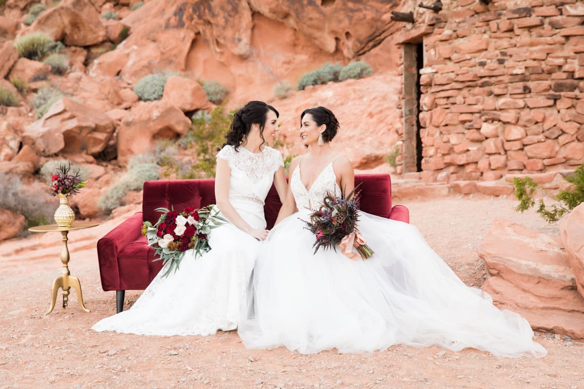 two brides in white sitting on red velvet couch in desert