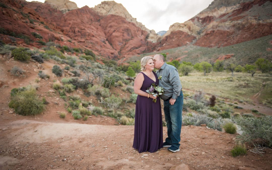 A husband kisses his wife on her cheek as they stand on a vista overlooking a valley and with mountains in the distance, after their vow renewal ceremony.