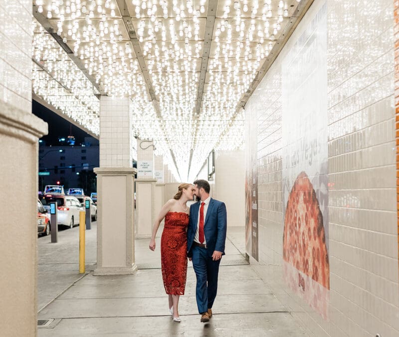 A couple in dressy clothes walks toward the camera leaning in for a kiss. They're outside in Las Vegas, walking under an awning covered in lights.