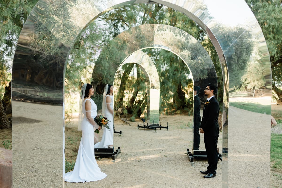 A bride and groom stand leaning against two sides of a reflective arch facing each other at GreenGale Farms in Las Vegas.